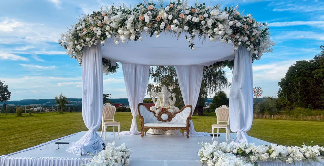 A large outdoor wedding altar with white fabric and flowers, featuring a white couch with a brown frame and a large white ...
